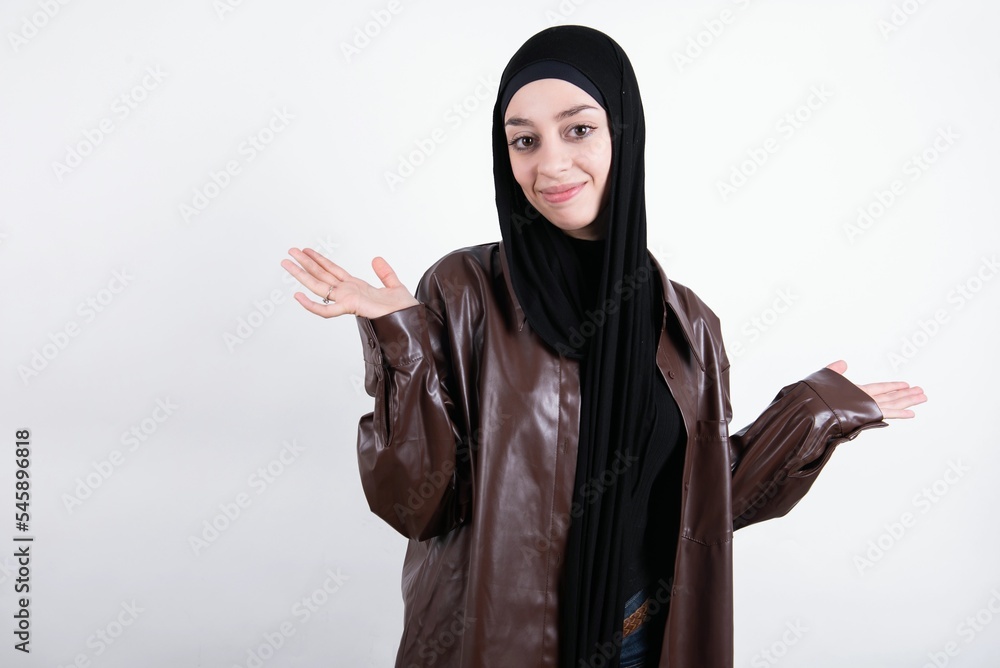 Puzzled and clueless young beautiful muslim woman wearing hijab and leather jacket over white with arms out, shrugging shoulders, saying: who cares, so what, I don't know. Negative human emotions. Puzzled and clueless young beautiful muslim woman wearing hijab and leather jacket over white with arms out, shrugging shoulders, saying: who cares, so what, I don't know. Negative human emotions.