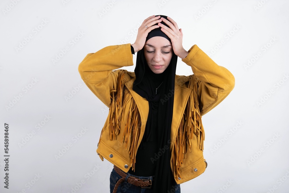 young beautiful muslim woman wearing hijab and yellow jacket over white background holding head with hands, suffering from severe headache, pressing fingers to temples young beautiful muslim woman wearing hijab and yellow jacket over white background holding head with hands, suffering from severe headache, pressing fingers to temples