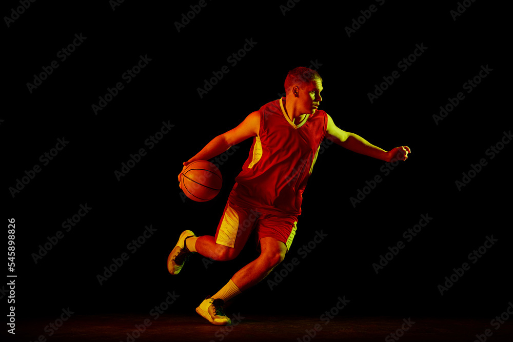 Dynamic portrait of young active athlete, male basketball player in sports uniform practicing with ball, jumping, dribbling isolated over dark background in neon light Dynamic portrait of young active athlete, male basketball player in sports uniform practicing with ball, jumping, dribbling isolated over dark background in neon light