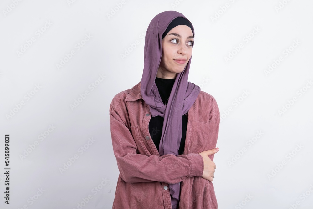 Pleased young beautiful muslim woman wearing hijab and pink jacket over white background keeps hands crossed over chest looks happily aside Pleased young beautiful muslim woman wearing hijab and pink jacket over white background keeps hands crossed over chest looks happily aside