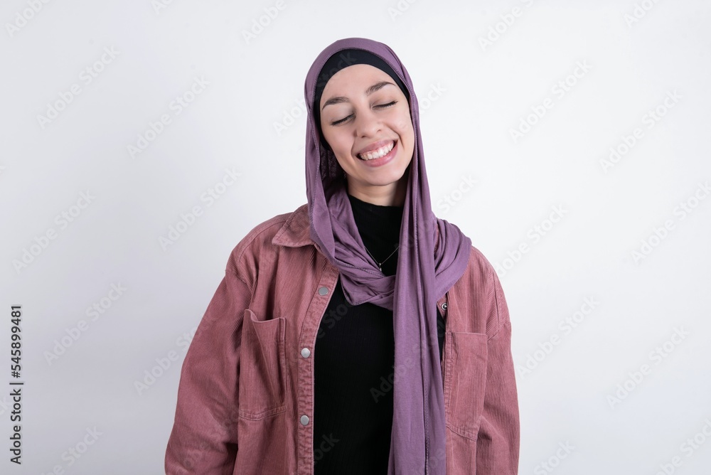 Positive young beautiful muslim woman wearing hijab and pink jacket over white background with overjoyed expression closes eyes and laughs shows white perfect teeth Positive young beautiful muslim woman wearing hijab and pink jacket over white background with overjoyed expression closes eyes and laughs shows white perfect teeth