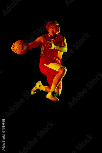 Active athletic male basketball player jumping with basketball ball isolated over dark background in red neon light. Concept of energy, professional sport, hobby.