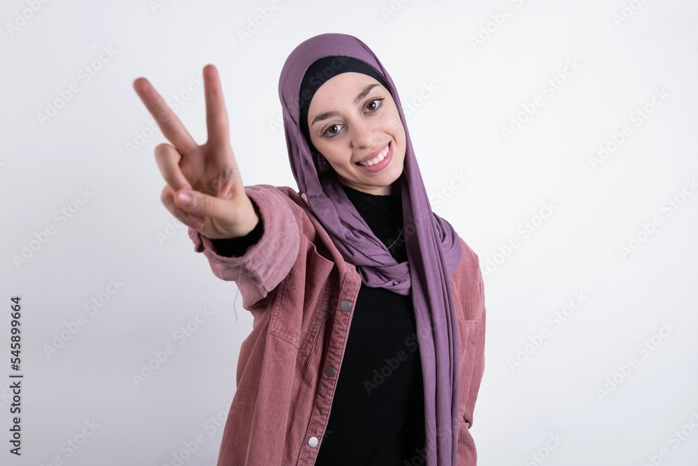 young beautiful muslim woman wearing hijab and pink jacket over white background directs fingers at camera selects someone. I recommend you. Best choice young beautiful muslim woman wearing hijab and pink jacket over white background directs fingers at camera selects someone. I recommend you. Best choice