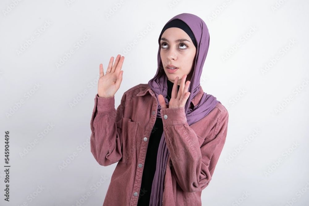 young beautiful muslim woman wearing hijab and pink jacket over white background keeps palms forward and looks with fright above on ceiling tries to defense herself from invisible danger opens mouth. young beautiful muslim woman wearing hijab and pink jacket over white background keeps palms forward and looks with fright above on ceiling tries to defense herself from invisible danger opens mouth.
