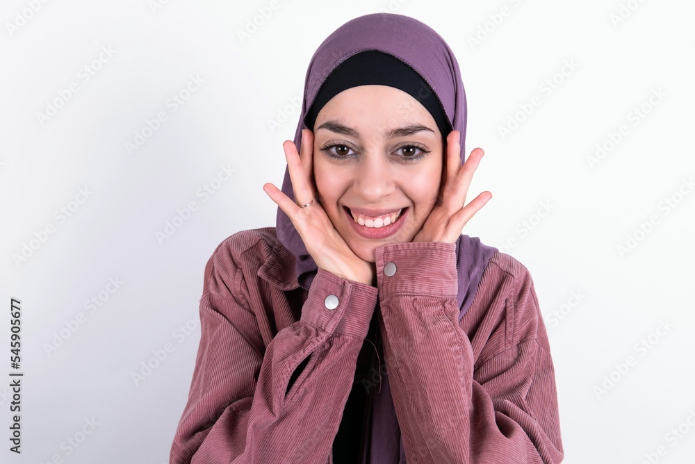 Happy young beautiful muslim woman wearing hijab against white background touches both cheeks gently, has tender smile, shows white teeth, gazes positively straightly at camera, Happy young beautiful muslim woman wearing hijab against white background touches both cheeks gently, has tender smile, shows white teeth, gazes positively straightly at camera,