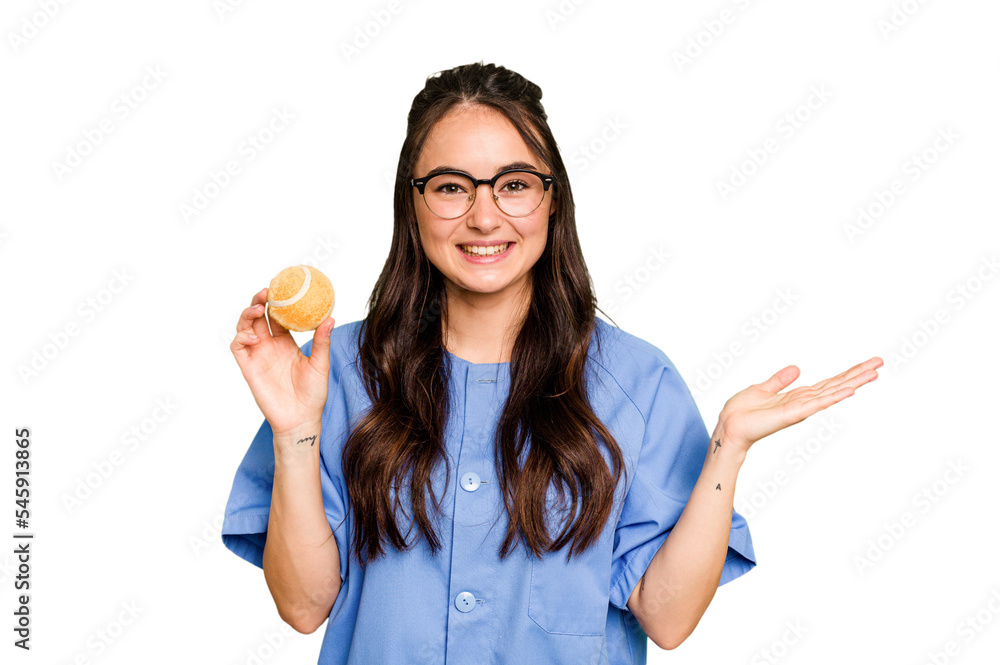 Young caucasian physiotherapist woman holding a tennis ball isolated isolated on green chroma background showing a copy space on a palm and holding another hand on waist. Young caucasian physiotherapist woman holding a tennis ball isolated isolated on green chroma background showing a copy space on a palm and holding another hand on waist.