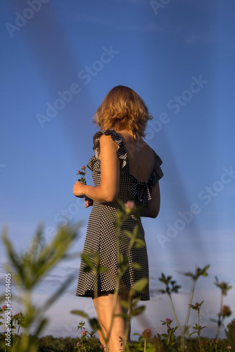 a woman in a black sundress with her hair down stands with her back in a field against a blue sky with grass in the foreground