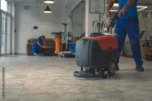 Cleaner cleans hard floor with scrubber machine while other cleaner cleans in the background