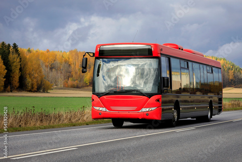 Bus with red front travelling on highway through rural scenery on an overcast day of autumn.