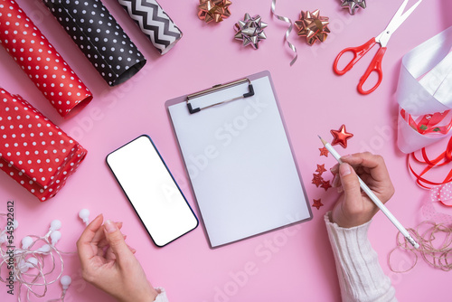 Blank sheet of paper, phone mock up and female hands wrapping christmas presents flat lay on pink background