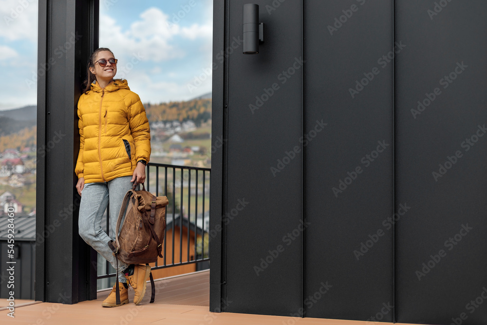 Young female in yellow down jacket with leather retro tourism backpack have a walk in nature background near eco houses Young female in yellow down jacket with leather retro tourism backpack have a walk in nature background near eco houses