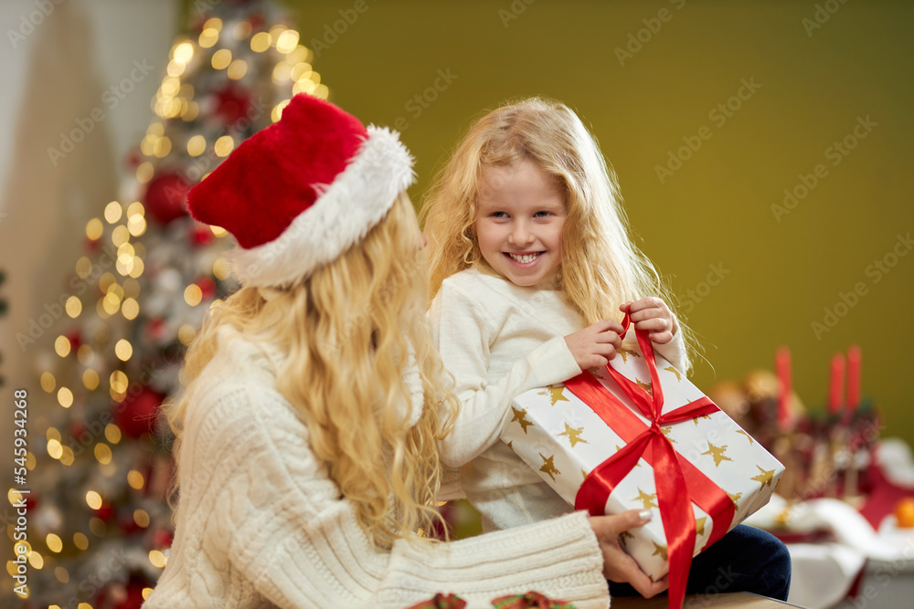 Side view of happy family sitting, posing indoors, at studio decorated with big christmas tree. Mother in christmas hat looking at daughter holding present. Concept of family. Side view of happy family sitting, posing indoors, at studio decorated with big christmas tree. Mother in christmas hat looking at daughter holding present. Concept of family.