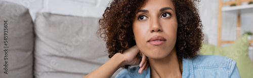 Canvas Print pensive african american woman with curly hair sitting near modern sofa in living room, banner