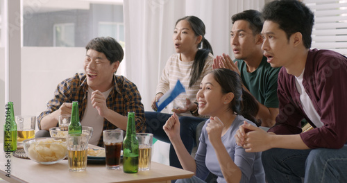 Group of young adult friend man and woman asia people sit at sofa couch joy chanting party fun game FIFA world cup live TV at home eat snack bowl drink beer bottle glass jump mad happy win exult face. photo