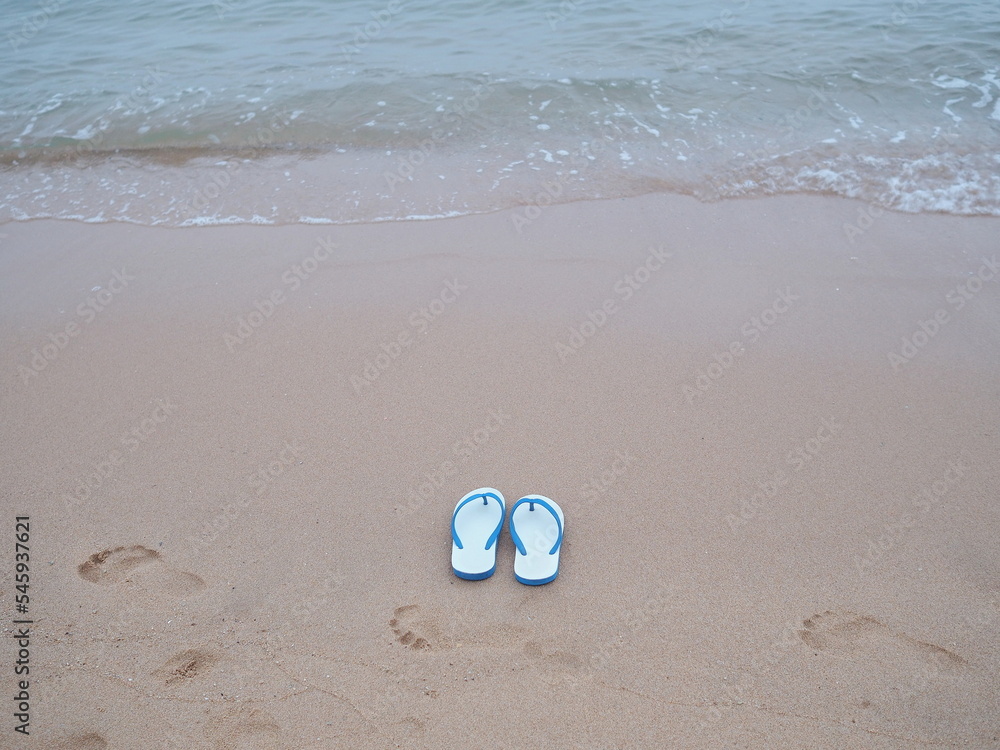 A pair of white  flip flops with blue straps rests on sandy beach with a small wave in front of the sea. Sponge shoes were removed and left in wet sand, and there were blurry images of human footprint