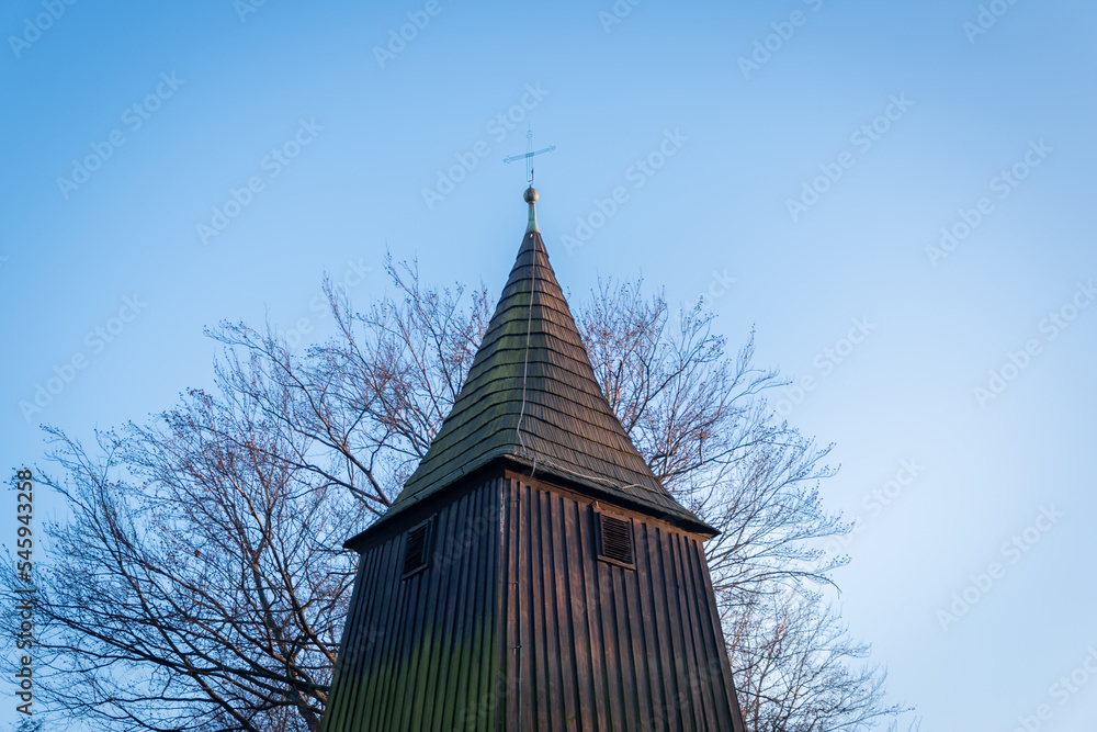 Metal cross on the peak of the belfry of the church of St. Michael the Archangel in Katowice, Silesia, Poland. Wooden temple from the beginning of XVI century. Leafless tree in the background.