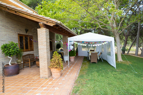 Big house with pines, prepared for a birthday party with the family, with a white tent, and some colour flags and decoration