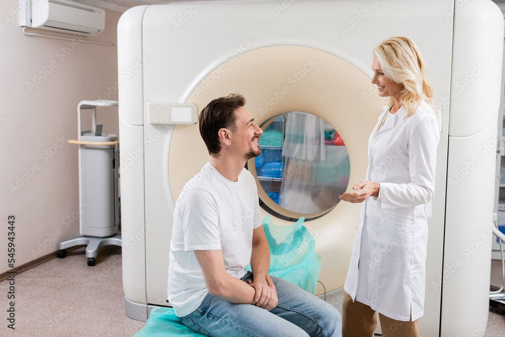 Obraz premium side view of blonde radiologist talking to smiling patient near computed tomography machine.