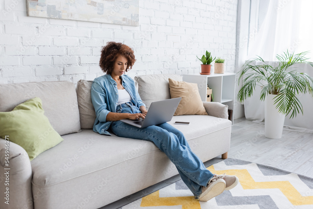 curly african american freelancer using laptop while sitting on couch and working from home. curly african american freelancer using laptop while sitting on couch and working from home.