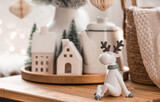 A bouquet of Christmas trees, reindeer, a blanket in a basket and Scandinavian white houses on a wooden table in the home interior of the living room. Cozy concept of festive decoration of the house.