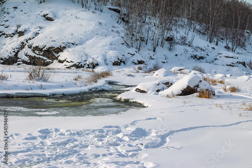 Winter landscape with a fast river with ice-free water, snow, dry grass and rocky shores
