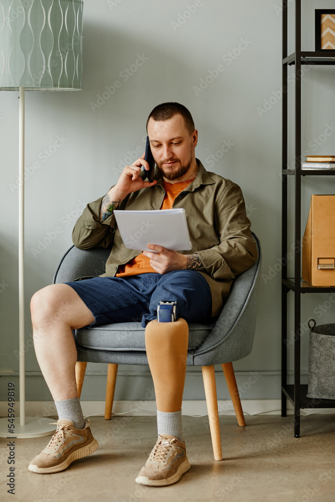 Full length portrait of man with prosthetic leg calling by phone while sitting in chair in home interior