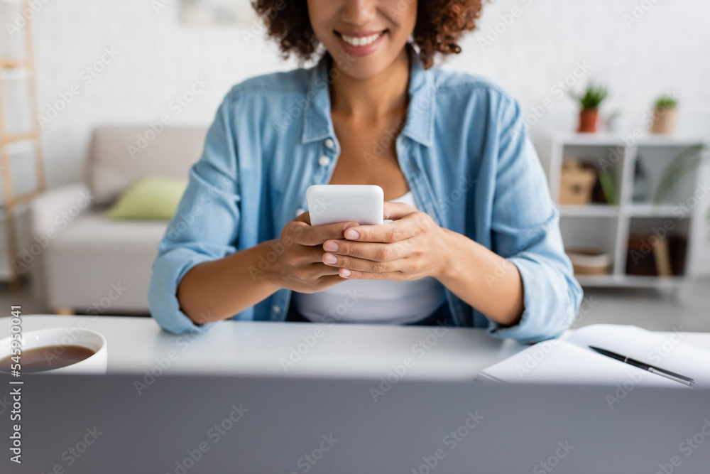 Cropped view of cheerful african american freelancer using smartphone near laptop and tea at home. Cropped view of cheerful african american freelancer using smartphone near laptop and tea at home.