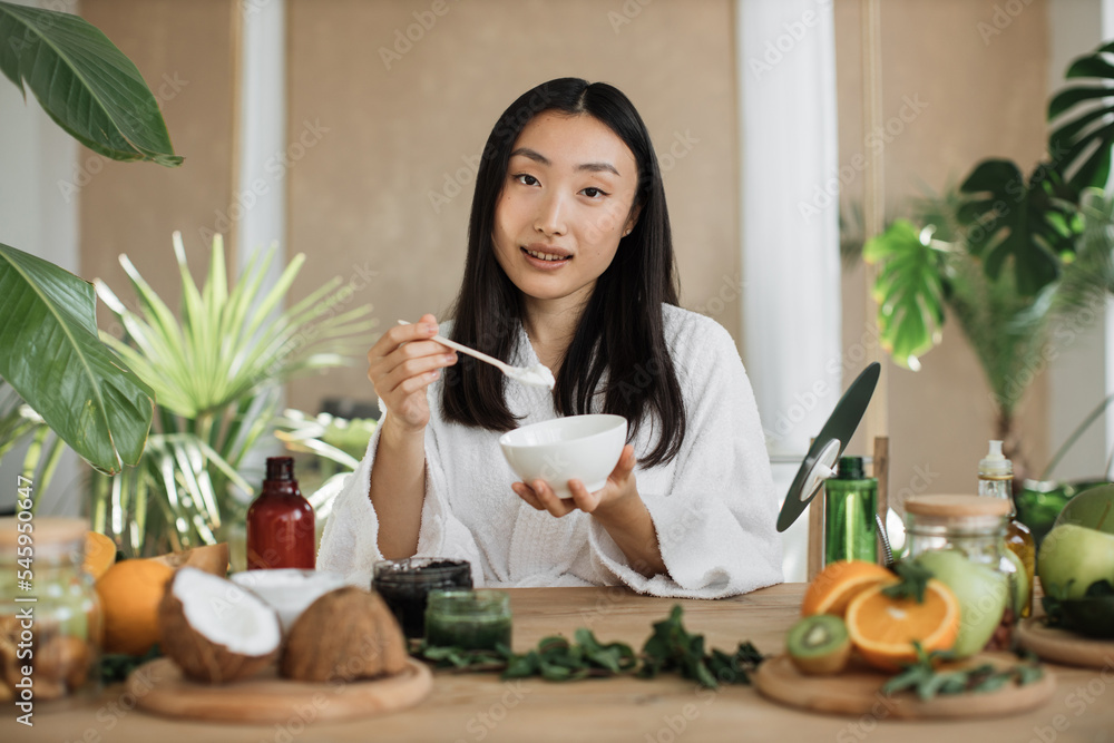 Beautiful healthy asian woman in white bathrobe sitting at wooden table with various ingredients preparing natural cosmetics at home, mixing the mass in a bowl while preparing cream. Skin care. Beautiful healthy asian woman in white bathrobe sitting at wooden table with various ingredients preparing natural cosmetics at home, mixing the mass in a bowl while preparing cream. Skin care.