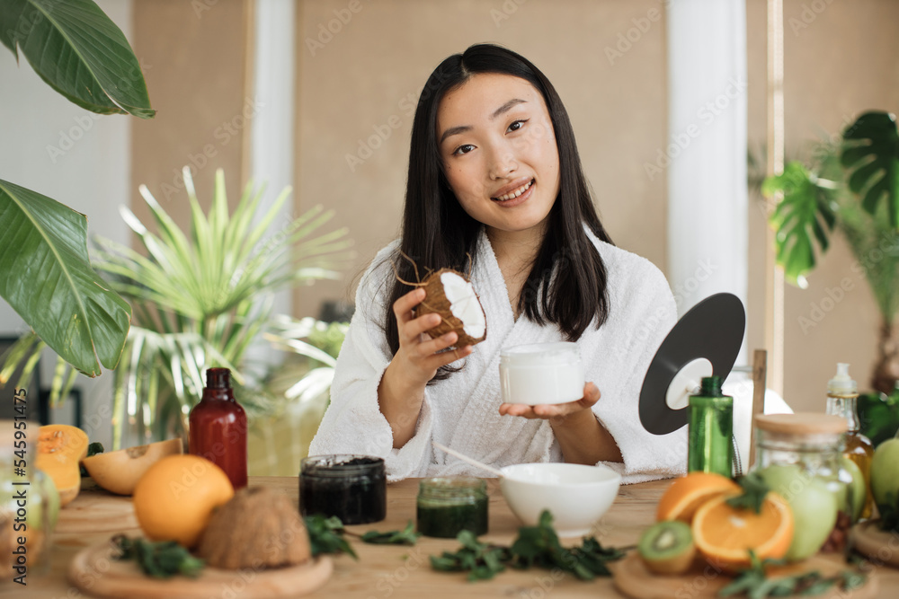 Young asian woman holding half of coconut and making homemade cream for healthy skin. A lot of ingredients for homemade cosmetics lying on wooden table. Young asian woman holding half of coconut and making homemade cream for healthy skin. A lot of ingredients for homemade cosmetics lying on wooden table.