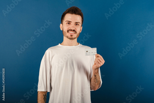 Portrait of young handsome man wearing white t-shirt posing isolated over blue background holding credit card and smiling with a confident smile showing teeth.