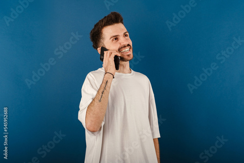 Happy casual man wearing t-shirt posing isolated over blue background talking on smart phone and smiling. Receiving good news during phone conversation.