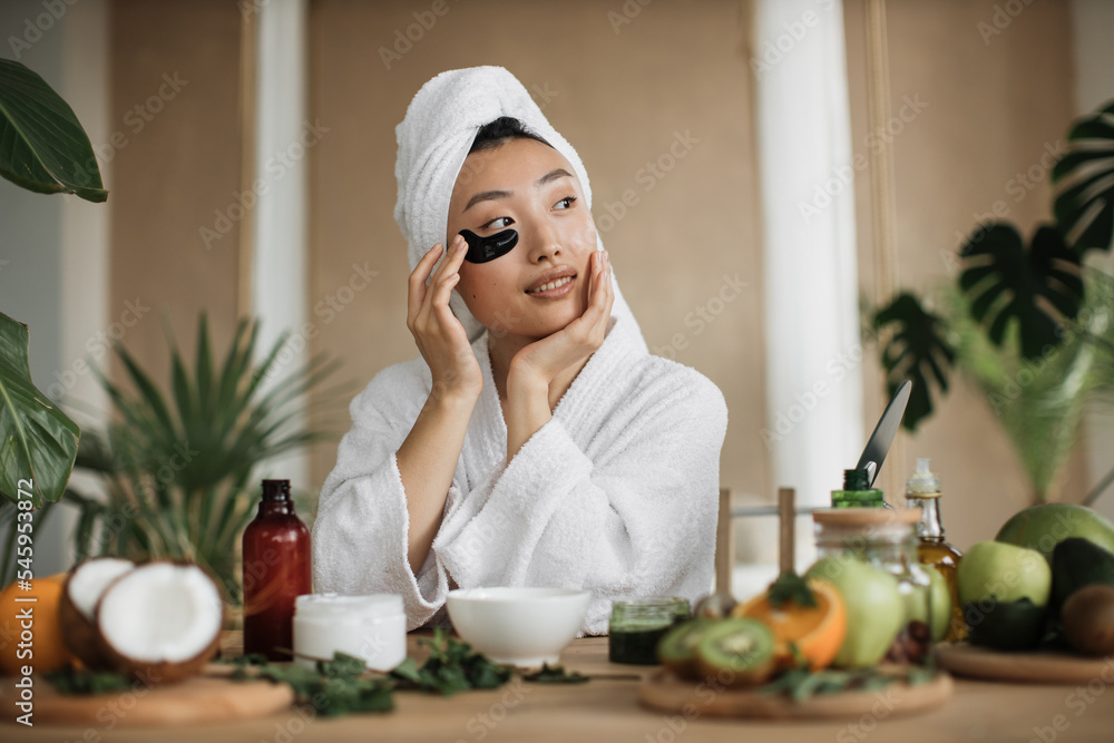 Pretty asian woman in towel and bathrobe sitting at table with fruits, vegetables for homemade cosmetics applying patches under eyes. Charming female making beauty procedures at exotic tropical home Pretty asian woman in towel and bathrobe sitting at table with fruits, vegetables for homemade cosmetics applying patches under eyes. Charming female making beauty procedures at exotic tropical home