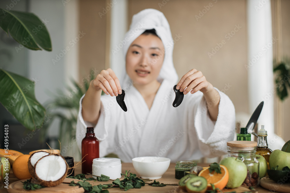 Focus on hands of smiling asian woman in towel on head and bathrobe sitting at table and holding collagen patches. Young female using homemade cosmetic for beauty routine at exotic studio. Focus on hands of smiling asian woman in towel on head and bathrobe sitting at table and holding collagen patches. Young female using homemade cosmetic for beauty routine at exotic studio.