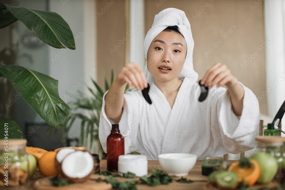 Smiling asian woman in towel on head and bathrobe sitting at table and holding collagen patches in hands. Young female using homemade cosmetic for beauty routine at exotic studio. Smiling asian woman in towel on head and bathrobe sitting at table and holding collagen patches in hands. Young female using homemade cosmetic for beauty routine at exotic studio.