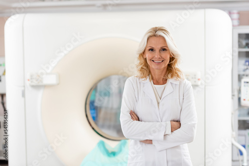 фотографія cheerful middle aged radiologist standing with crossed arms near computed tomography scanner