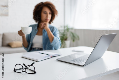 laptop and stationery on desk near blurred african american freelancer with cup.