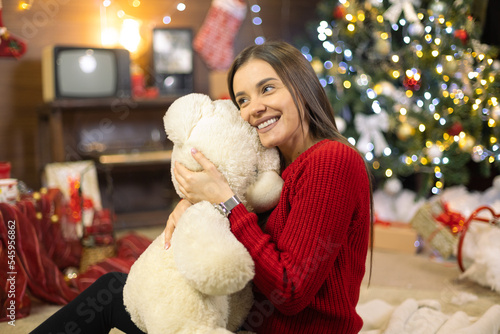 Beautiful pregnant girl in front of Christmas tree and New Year decorations, sitting alone with a stuffed toy, a teddy bear, smiling, enjoying the background are gifts old TV and old furniture