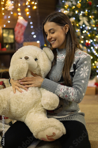 Beautiful pregnant girl in front of Christmas tree and New Year decorations, sitting alone with a stuffed toy, a teddy bear, smiling, enjoying the background are gifts old TV and old furniture
