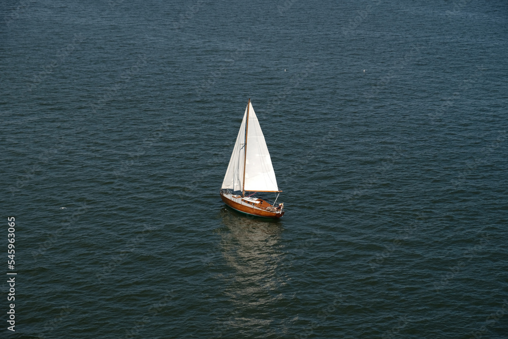 Fototapeta premium Aerial view of wooden old yacht with sail against backdrop of blue water with waves and ripples. Lonely white sail is sailing. Perfect content for posters or advertising banners, creative projects.