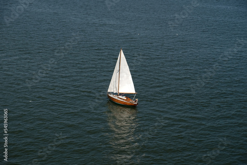 Aerial view of wooden old yacht with sail against backdrop of blue water with waves and ripples. Lonely white sail is sailing. Perfect content for posters or advertising banners, creative projects.