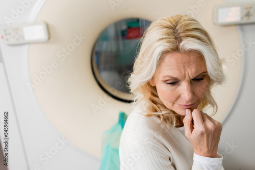 nervous middle aged woman holding hand near face near ct scanner on blurred background.