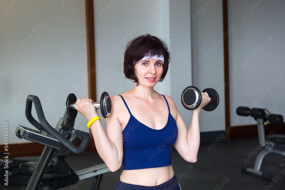 Fototapeta premium Sports woman lifting dumbbells in gym with large window.