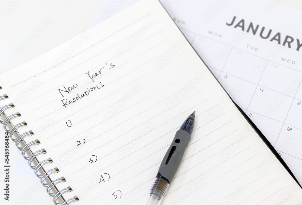 Notepad with handwritten New Year’s resolutions list and pen on top of January calendar in the background, selective focus Notepad with handwritten New Year’s resolutions list and pen on top of January calendar in the background, selective focus
