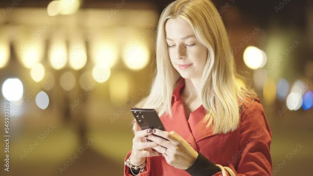 Attractive blonde woman in red coat smile at smartphone at night in bright city 4K Attractive blonde woman in red coat smile at smartphone at night in bright city 4K