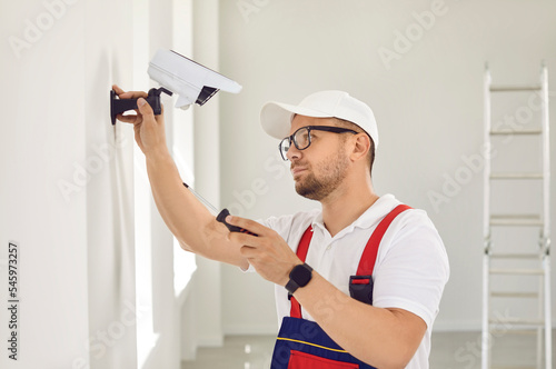 A Caucasian male repairman in uniform and glasses holds a screwdriver and installs video surveillance equipment on the wall inside the room. The concept of security and protection.