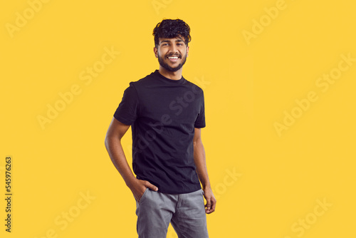 Young cheerful attractive South Asian man with dark curly hair posing with smile in casual clothes holding hand in pocket of trousers stands on solid yellow background in studio. Ethnic Indian human