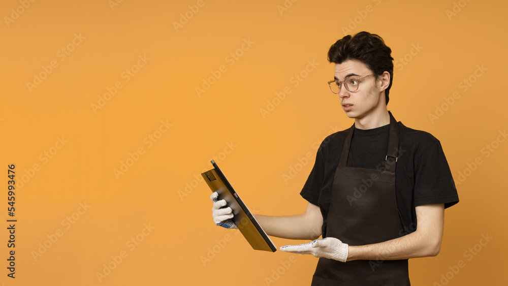 young man in a black t-shirt and apron in work gloves with a tablet for papers in his hands is perplexed and looks away at copy space