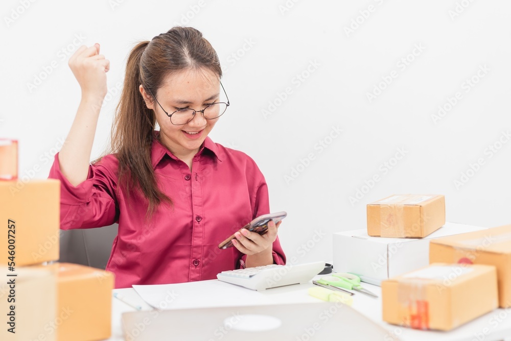 Online selling concept, happy and smiling Asian woman, reading message on smartphone screen, desk with laptop Cardboard boxes and calculators. Online selling concept, happy and smiling Asian woman, reading message on smartphone screen, desk with laptop Cardboard boxes and calculators.