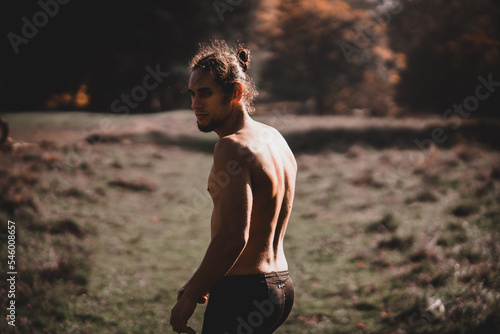 Adult man with a shirtless ponytail looking down at the ground in a forest