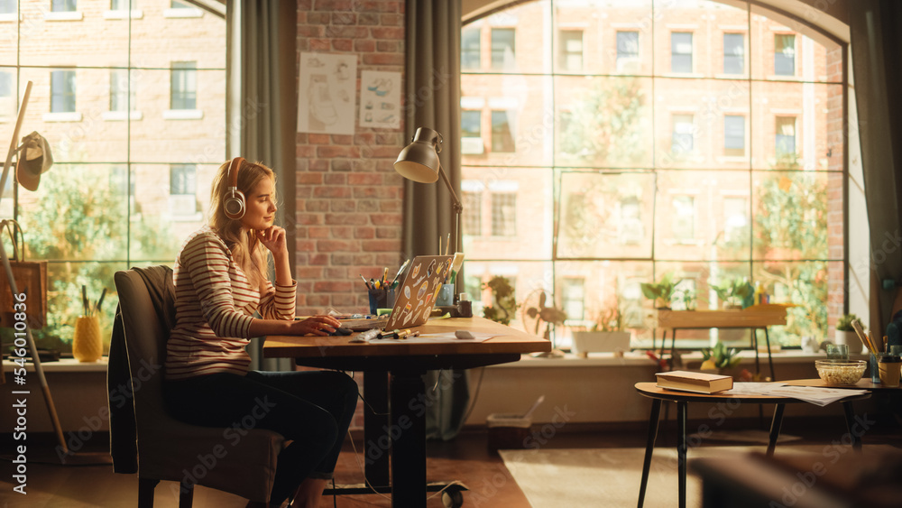 Young Pretty Woman with Blond Hair Wearing Headphones and Working from Home on Laptop Computer in Stylish Loft Apartment. Creative Female Smiling, Checking Social Media, Browsing Internet. Young Pretty Woman with Blond Hair Wearing Headphones and Working from Home on Laptop Computer in Stylish Loft Apartment. Creative Female Smiling, Checking Social Media, Browsing Internet.